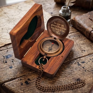 May include: A wooden compass in a hinged box, open to reveal the compass face and a message on the lid. The compass has a brass finish and a chain. The box is made of dark wood and lined with green felt. The text on the lid reads: "So you can always find your way back to me, this."