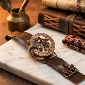 Op de afbeelding: Een koperen zonnewijzerhorloge met een ronde wijzerplaat voorzien van Romeinse cijfers en een centrale gnomon, bevestigd aan een brede bruine leren band met dubbele metalen gespen. De wijzerplaat is gegraveerd met de tekst West London.