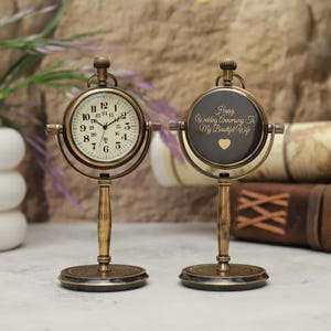 May include: Two antique-style brass desk clocks on circular bases. The left clock features a cream face with black numerals and a 24-hour scale. The right clock displays the text "Happy Wedding Anniversary To My Beautiful Wife" with a heart icon.