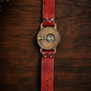 May include: A wristwatch with a red leather strap and a brass-toned circular face. The dial features engraved markings, numbers, and a central compass-like needle with a small green accent. The strap is secured with metal rivets and a buckle.