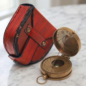 May include: A vintage brass sundial compass with an open lid, displaying etched markings and a green gem. Beside it is a red leather case with a flap and brass studs. The compass is on a marble surface.