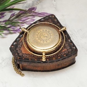 May include: A circular brass sundial compass with Roman numerals and engraved markings sits atop a triangular, dark brown leather case featuring decorative stitching and embossed patterns. A gold-toned metal chain is attached to the side.