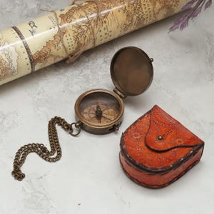 Può includere: Una bussola da tasca in ottone con coperchio aperto e catena in metallo, accompagnata da una piccola custodia in pelle arancione goffrata con un motivo a rosa dei venti. La bussola presenta un quadrante circolare con punti cardinali.