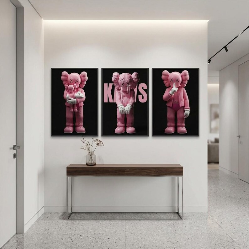 Pu&ograve; includere: Tre stampe artistiche incorniciate con personaggi rosa dei cartoni animati su sfondo nero. Una figura tiene una figura pi&ugrave; piccola, un'altra ha la parola "KAWS" in rosa e la terza ha un dito sulle labbra. Sotto c'&egrave; una consolle in legno con un vaso di fiori.