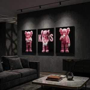 Pu&ograve; includere: Tre opere d'arte incorniciate con personaggi di cartoni animati rosa con occhi e mani a forma di X. Le figure sono su sfondo nero, con la parola "KAWS" in rosa su una di esse. L'arte &egrave; esposta in un moderno soggiorno.