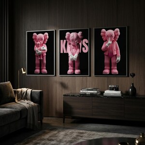 Pu&ograve; includere: Tre stampe artistiche incorniciate con personaggi di cartoni animati rosa con occhi a forma di X e mani guantate. Una figura tiene una figura pi&ugrave; piccola, un'altra ha la parola "KAWS" in rosa e la terza ha un dito sulle labbra. Lo sfondo &egrave; nero.