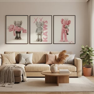 May include: Three framed KAWS art prints. The prints feature cartoon figures in various poses, with pink and gray color schemes. The artwork is displayed above a beige sectional sofa with decorative pillows and a round wooden coffee table.