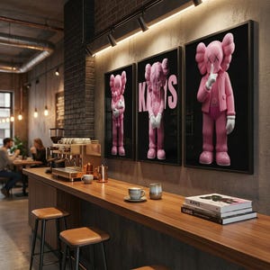 Pu&ograve; includere: Tre stampe artistiche KAWS rosa incorniciate appese a una parete in un bar. Le stampe presentano il personaggio KAWS in varie pose su sfondo nero. Un bancone in legno con sgabelli, tazze di caff&egrave; e libri &egrave; in primo piano.