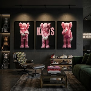 Pu&ograve; includere: Un moderno soggiorno con tre opere d'arte KAWS rosa incorniciate. La stanza comprende una poltrona in pelle con stampa mimetica, un tavolino con libri e un'esposizione di sneakers. L'estetica generale &egrave; contemporanea ed elegante.