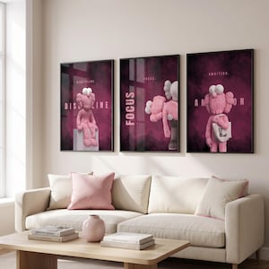 Peut inclure: Trois cadres muraux représentant des personnages de dessin animé roses aux yeux en forme de X sur un fond fumé magenta foncé. L'affiche de gauche affiche le mot DISCIPLINE, celle du centre FOCUS et celle de droite AMBITION.
