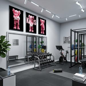 Pu&ograve; includere: Interno di palestra moderna con pavimenti neri, attrezzature per l'allenamento e tre stampe rosa incorniciate di KAWS sulla parete. Le stampe presentano una figura di cartone animato rosa. Il testo "MIND OVER MATTER" &egrave; su un cartello.