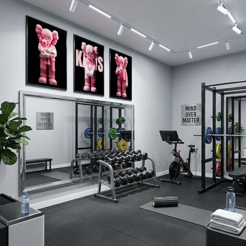 Pu&ograve; includere: Interno di palestra moderna con pavimenti neri, attrezzature per l'allenamento e tre stampe rosa incorniciate di KAWS sulla parete. Le stampe presentano una figura di cartone animato rosa. Il testo "MIND OVER MATTER" &egrave; su un cartello.