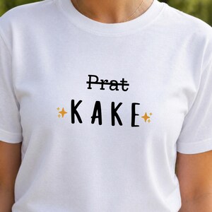 Puede incluir: Camiseta blanca con el texto "Prat KAKE" en fuente negra. La palabra "Prat" está tachada. Gráficos de estrellas doradas a ambos lados de la palabra "KAKE".