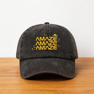 Può includere: Un cappellino da baseball nero con la parola "AMAZE" in giallo, impilata tre volte con un design a pianeta stilizzato. Il cappellino è realizzato in un materiale morbido e usurato.