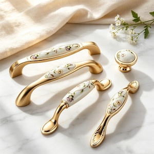 Könnte beinhalten: Eine Sammlung goldfarbener Schrankbeschläge, darunter Griffe und ein Knauf. Die Beschläge weisen weiße Keramikeinsätze mit zarten Blumenmustern in Lila, Gelb und Grün auf. Die Hardware ist auf einer Marmoroberfläche angeordnet.
