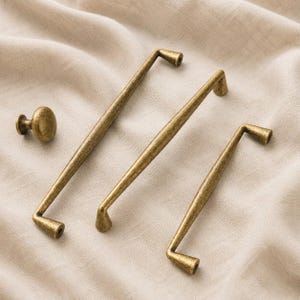 Könnte beinhalten: Ein Satz antiker Messing-Schrankbeschläge, bestehend aus drei Griffen und einem Knauf. Die Griffe haben ein schlankes, abgewinkeltes Design, und der Knauf ist rund. Die Beschläge sind auf einem neutralen Stoffhintergrund dargestellt.