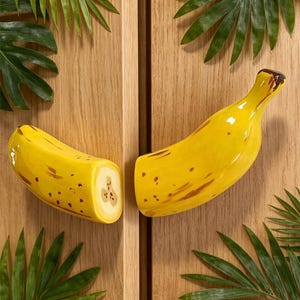 Könnte beinhalten: Zwei gelbe, bananenförmige Schrankgriffe aus Keramik mit braunen Flecken und einem braunen Stiel. Sie sind vor einem hellen Holzgrund platziert, mit grünen Palmenblättern in den Ecken.