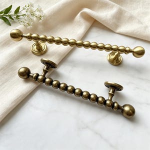May include: Two antique brass door pulls with a beaded design. Each pull features a series of small spheres connected in a line, with larger spheres at each end. The pulls are mounted on round bases.