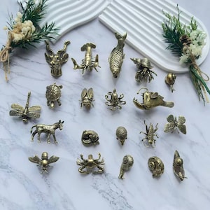 Könnte beinhalten: Eine Sammlung kleiner, antiker Messingfiguren, die verschiedene Tiere und Insekten darstellen. Die Auswahl umfasst einen Fisch, eine Krabbe, eine Libelle, einen Schmetterling und einen Esel. Die Figuren sind auf einer Marmoroberfläche angeordnet, mit dekorativem Grün im Hintergrund.