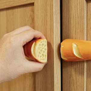 Könnte beinhalten: Zwei keramische Schrankknöpfe in Baguette-Form. Einer wird von einer Hand gehalten, die die Kruste und das Innere des Brotes zeigt. Die Knöpfe sind in Braun- und Gelbtönen bemalt und verleihen der Küchen- oder Schrankdekoration eine verspielte Note.