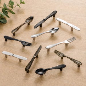 Könnte beinhalten: Eine Sammlung dekorativer Schrankgriffe in Form von Miniatur-Besteck. Die Griffe umfassen Löffel, Gabeln und Messer in verschiedenen Ausführungen, darunter Schwarz, Silber und Antik-Bronze. Sie sind auf einer hellen Holzoberfläche angeordnet.