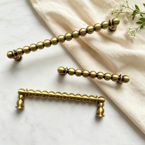 May include: Three antique brass cabinet pulls with a beaded design. The pulls are in varying sizes and are arranged on a white surface with a beige cloth and small white flowers.