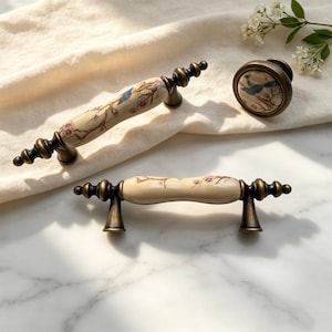 Op de afbeelding: Een set van drie meubelbeslagstukken in antieke stijl. De set bevat twee ladetrekker en een knop. Elk stuk heeft een crèmekleurig keramisch midden met een vogel- en bloemmotief, geplaatst op een bronzen metalen basis.