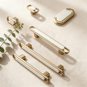 Könnte beinhalten: Eine Sammlung goldfarbener Schrankbeschläge mit weißen Keramikeinsätzen. Das Set umfasst verschiedene Größen von Griffen und Knöpfen, die ein modernes Design zeigen. Die Beschläge sind auf einer neutralen Oberfläche angeordnet, was den Kontrast zwischen Gold und Weiß hervorhebt.