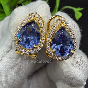 May include: A pair of gold-toned earrings featuring large, pear-shaped blue gemstones at the centre. Each stone is surrounded by a halo of small, clear, sparkling stones set in a gold-coloured metal frame. The earrings have a secure clasp mechanism.