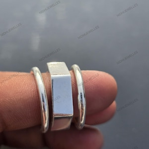 Könnte beinhalten: Ein Satz von drei silbernen Ringen. Der zentrale Ring ist rechteckig, flankiert von zwei abgerundeten Bändern. Die Ringe werden auf einem Finger präsentiert, um ihr Design und ihren metallischen Glanz zu zeigen. Der Schmuck ist schlicht und elegant.