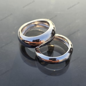 May include: Two polished silver rings with a rounded design are displayed on a reflective surface. The rings are close together, with one slightly larger than the other. The rings' smooth surfaces reflect the surrounding environment.