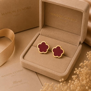 Peut inclure: Une paire de boucles d'oreilles dorées en forme de fleur à cinq pétales avec un centre bordeaux profond. Les boucles d'oreilles sont présentées dans un écrin en velours beige avec le texte MAISON CENDRE PARIS imprimé sur le couvercle et l'intérieur.
