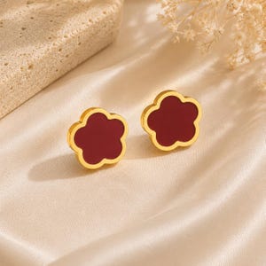 Puede incluir: Un par de pendientes de botón con forma de flor de cinco pétalos, con un borde de metal dorado y un centro de color rojo oscuro sólido. Los pendientes tienen un diseño suave y redondeado con un acabado pulido, ofreciendo una estética floral.