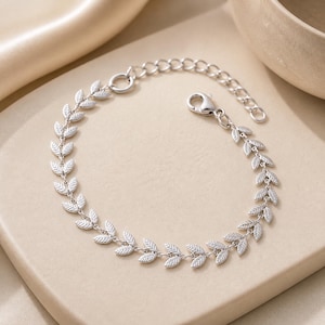 Peut inclure: Bracelet argenté orné d'un motif de feuilles délicates. Le bracelet est doté d'une chaîne réglable et d'un fermoir mousqueton. Les feuilles sont espacées de manière uniforme, créant un motif symétrique. Le bijou est présenté sur une surface neutre.