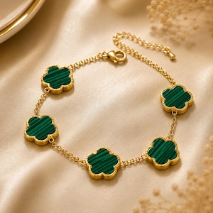 Può includere: Un bracciale a catena color oro con cinque ciondoli a forma di fiore distanziati uniformemente. Ogni ciondolo presenta un inserto a righe verde scuro con un bordo color oro liscio e lucido. Il bracciale include una catena sottile e una chiusura a moschettone.