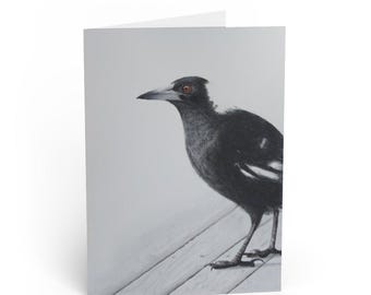 Op de afbeelding: Wenskaart met een gedetailleerde illustratie van een ekster. De vogel is weergegeven in grijstinten, met zwart-witte veren en een opvallend oranje oog. De ekster staat op een houten oppervlak tegen een witte achtergrond.