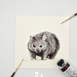 Puede incluir: Una hoja de papel cuadrada con una ilustración detallada en acuarela de un wombat gris de pie sobre hierba verde claro. Se incluyen dos pinceles finos con mangos de madera y un tubo de pintura de acuarela Daniel Smith Sombra Tostada.