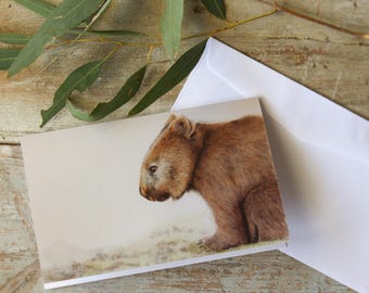 May include: A rectangular greeting card featuring a detailed illustration of a brown wombat in profile, positioned on a light-colored ground. The card is accompanied by a plain white envelope, both resting on a weathered wooden surface.