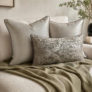 May include: Three decorative cushions in silver and grey tones are arranged on a cream sofa. Two square cushions have a textured finish with black piping. A rectangular cushion displays an ornate pattern. An olive green throw blanket adds texture.