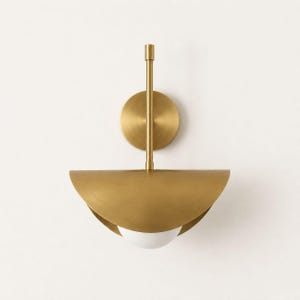 Puede incluir: Aplique de pared moderno con acabado en latón. La luminaria presenta una placa trasera circular, una varilla vertical y una pantalla semicircular. Una bombilla esférica blanca es visible dentro de la pantalla. Diseñado para iluminación interior.