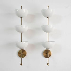 May include: Two matching wall sconces with a mid-century modern design. Each sconce features three white, bowl-shaped shades stacked vertically on a brass rod. The sconces have a brass base and a decorative brass finial.