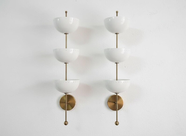 May include: Two matching wall sconces with a mid-century modern design. Each sconce features three white, bowl-shaped shades stacked vertically on a brass rod. The sconces have a brass base and a decorative brass finial.