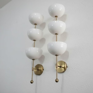 May include: Two wall sconces with a mid-century modern design. Each sconce features three white, bowl-shaped shades stacked vertically, connected by a brass rod. The sconces are mounted on brass circular backplates.