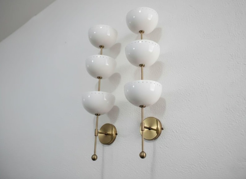 May include: Two wall sconces with a mid-century modern design. Each sconce features three white, bowl-shaped shades stacked vertically, connected by a brass rod. The sconces are mounted on brass circular backplates.