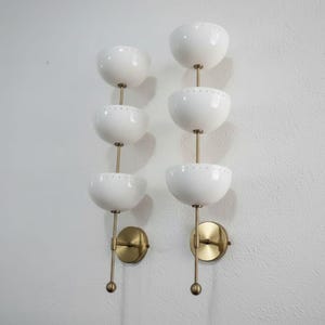 May include: Two wall sconces with a mid-century modern design. Each sconce features three white, bowl-shaped shades stacked vertically, connected by a brass rod. The sconces have brass mounting plates and are mounted on a white wall.