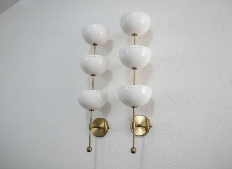May include: Two wall sconces with a mid-century modern design. Each sconce features three white, bowl-shaped shades stacked vertically, connected by a brass rod. The sconces have brass mounting plates and are mounted on a white wall.