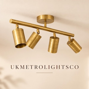 Op de afbeelding: Een messing plafondlamp met een horizontale stang en vier verstelbare cilindrische spots. De armatuur heeft een ronde montageplaat en een geborstelde metalen afwerking. De tekst UKMETROLIGHTSCO staat hieronder vermeld.