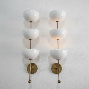 May include: Two matching wall sconces with a mid-century modern design. Each sconce features three white, bowl-shaped shades stacked vertically on a brass stem. The shades have small, decorative perforations. The sconces are mounted on brass backplates.
