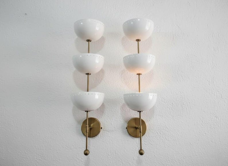 May include: Two matching wall sconces with a mid-century modern design. Each sconce features three white, bowl-shaped shades stacked vertically on a brass stem. The shades have small, decorative perforations. The sconces are mounted on brass backplates.