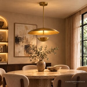 May include: A brass pendant light fixture featuring a large, shallow, saucer-shaped shade positioned above a smaller, inverted bowl-shaped shade, suspended by a thin vertical rod from a circular ceiling mount, with the text UKMETROLIGHTSCO.